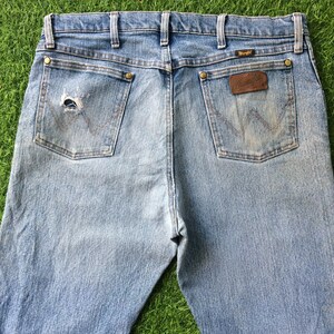 May include: A pair of light blue denim jeans with a distressed back pocket and a brown leather Wrangler label.