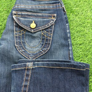 May include: Dark wash denim jeans with a classic five-pocket design. Features a decorative back pocket with gold stitching and a gold button. A brown leather patch is visible on the back waistband.
