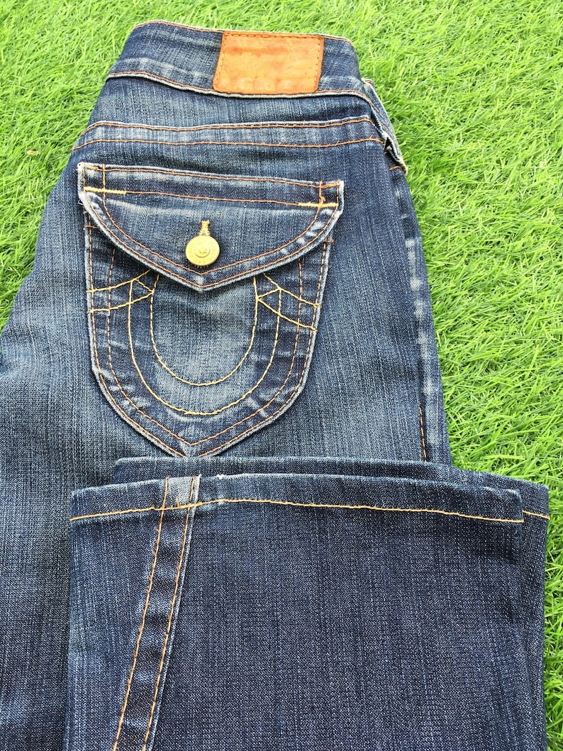 May include: Dark wash denim jeans with a classic five-pocket design. Features a decorative back pocket with gold stitching and a gold button. A brown leather patch is visible on the back waistband.