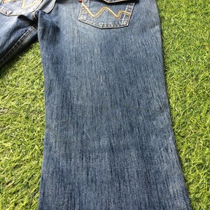 May include: Blue denim jeans with decorative stitching on the back pockets. The jeans have a classic five-pocket design, a leather patch, and a straight leg. The jeans are displayed on a green surface.