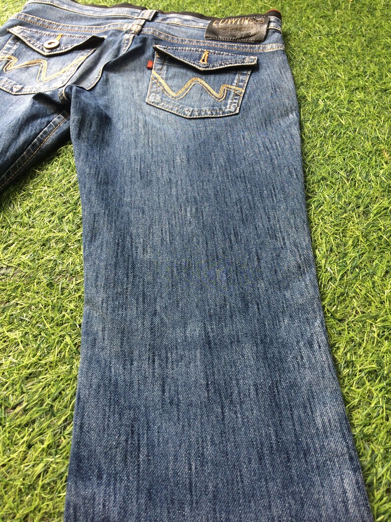 May include: Blue denim jeans with decorative stitching on the back pockets. The jeans have a classic five-pocket design, a leather patch, and a straight leg. The jeans are displayed on a green surface.