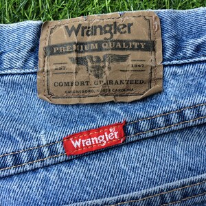 Pode incluir: Um close-up de um par de cal&ccedil;as de ganga azuis com uma etiqueta Wrangler de couro castanho que diz "Premium Quality, Est. 1947, Comfort Guaranteed, Greensboro, North Carolina". H&aacute; tamb&eacute;m uma etiqueta Wrangler vermelha costurada nas cal&ccedil;as.