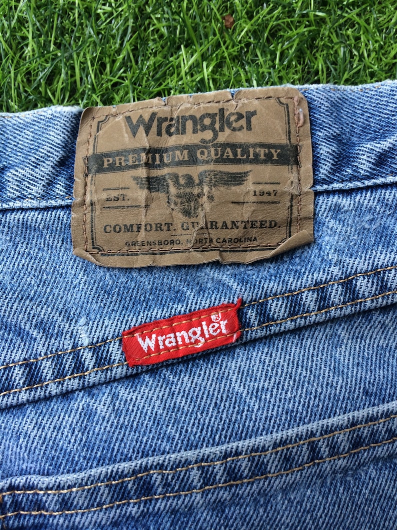 Pode incluir: Um close-up de um par de cal&ccedil;as de ganga azuis com uma etiqueta Wrangler de couro castanho que diz "Premium Quality, Est. 1947, Comfort Guaranteed, Greensboro, North Carolina". H&aacute; tamb&eacute;m uma etiqueta Wrangler vermelha costurada nas cal&ccedil;as.