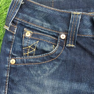May include: Close-up of dark blue denim jeans. The jeans feature a front pocket with decorative stitching, metal rivets, and contrasting yellow stitching. The fabric shows a worn, textured appearance.