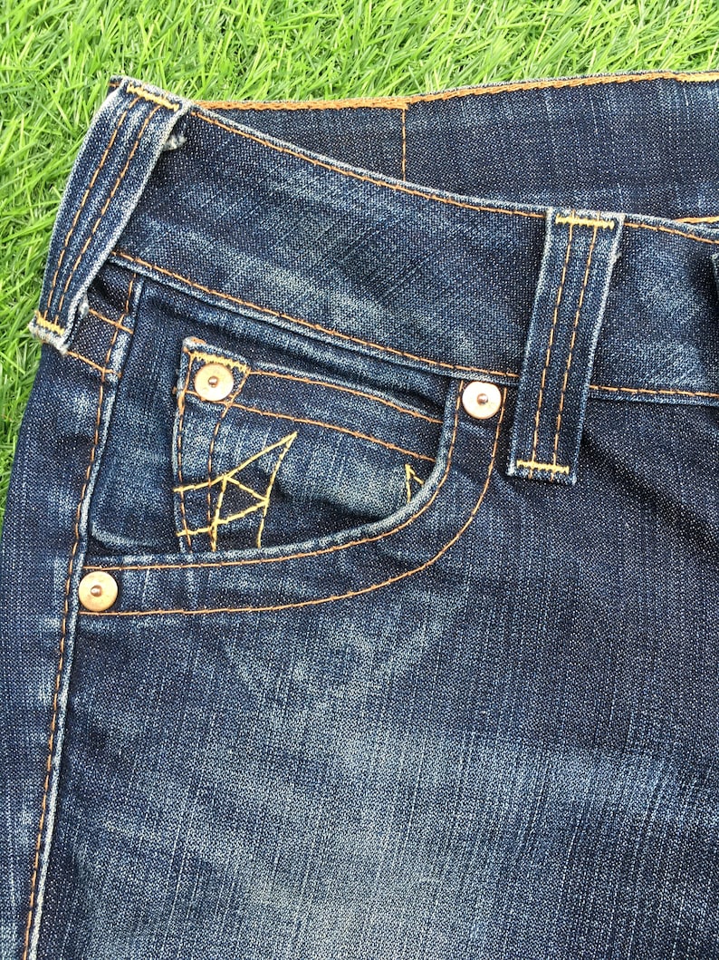 May include: Close-up of dark blue denim jeans. The jeans feature a front pocket with decorative stitching, metal rivets, and contrasting yellow stitching. The fabric shows a worn, textured appearance.