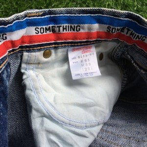 May include: Blue denim jeans with a red, white and blue waistband. The waistband has the text "SOMETHING" on it. The jeans have a white tag with the text "Something Ones" and the size information "LOT 9213-96, W 61, H 88 (28)".