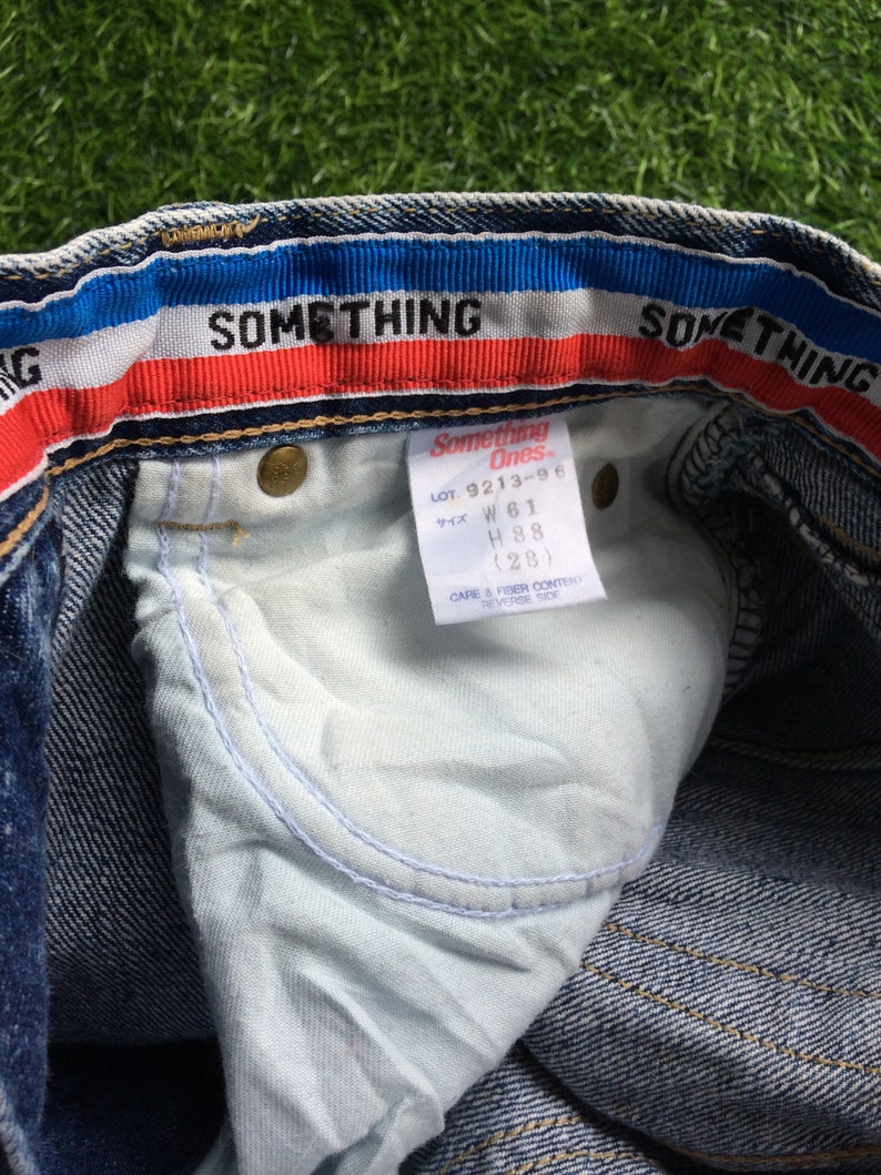 May include: Blue denim jeans with a red, white and blue waistband. The waistband has the text "SOMETHING" on it. The jeans have a white tag with the text "Something Ones" and the size information "LOT 9213-96, W 61, H 88 (28)".