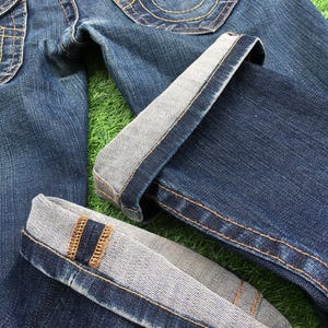 May include: Close-up of blue denim jeans with folded cuffs. The jeans feature gold stitching, a button closure, and a pocket with a decorative stitch design. The cuffs reveal a lighter fabric.