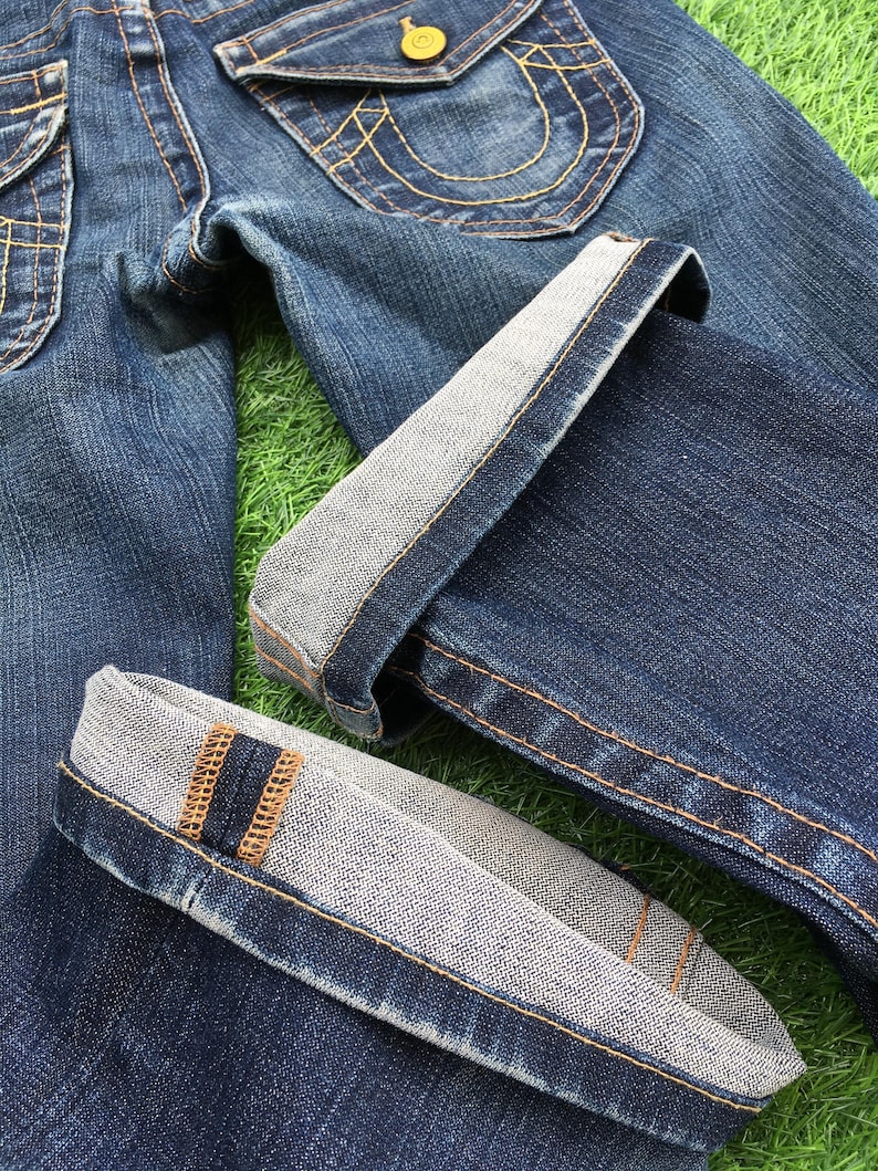 May include: Close-up of blue denim jeans with folded cuffs. The jeans feature gold stitching, a button closure, and a pocket with a decorative stitch design. The cuffs reveal a lighter fabric.