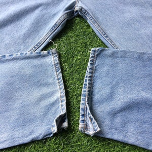May include: A pair of light blue denim jeans with the seams ripped at the bottom of the legs. The jeans are laid out on a green grassy surface.