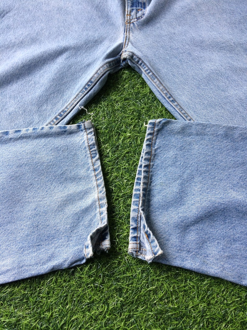 May include: A pair of light blue denim jeans with the seams ripped at the bottom of the legs. The jeans are laid out on a green grassy surface.