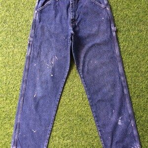 May include: A pair of blue denim carpenter jeans with white paint splatters. The jeans have a classic five-pocket style and hammer loops.