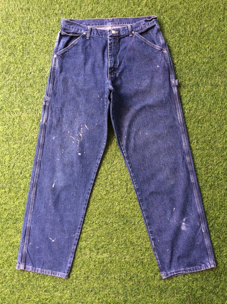 May include: A pair of blue denim carpenter jeans with white paint splatters. The jeans have a classic five-pocket style and hammer loops.