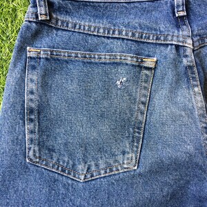 May include: Close-up of the back pocket of a pair of blue denim jeans. The pocket has light blue stitching detail around the edges.