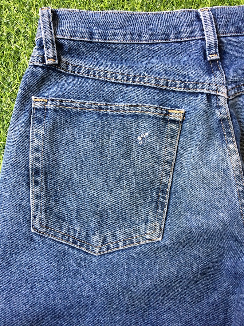 May include: Close-up of the back pocket of a pair of blue denim jeans. The pocket has light blue stitching detail around the edges.