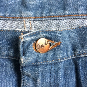 May include: Close-up of a blue denim jean with a copper-colored button. The button has a small, raised design.
