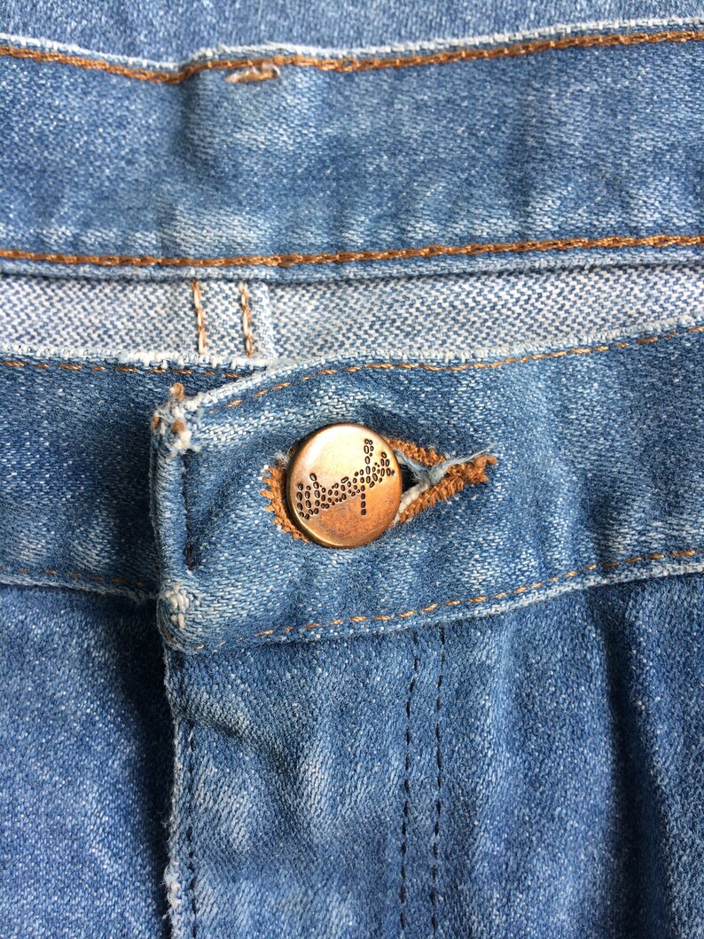 May include: Close-up of a blue denim jean with a copper-colored button. The button has a small, raised design.