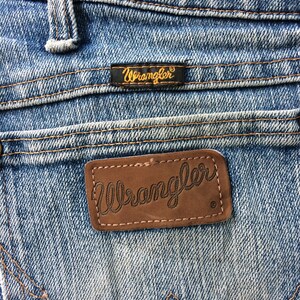 May include: Close-up of a pair of blue denim jeans with a brown leather Wrangler label. The label has the word "Wrangler" stitched in white thread.