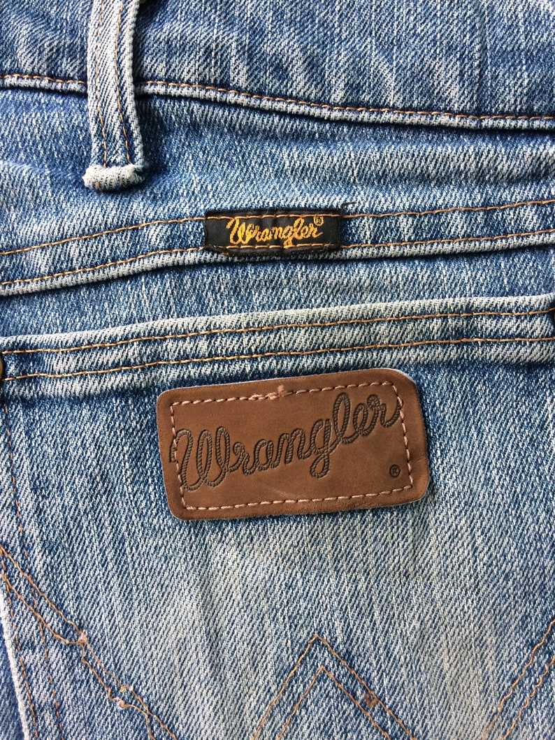 May include: Close-up of a pair of blue denim jeans with a brown leather Wrangler label. The label has the word "Wrangler" stitched in white thread.