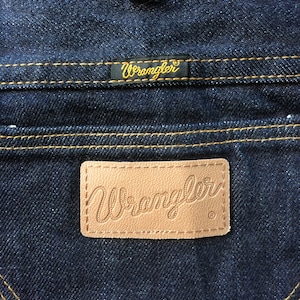 Peut inclure: Gros plan sur une paire de jeans en denim bleu fonc&eacute; avec un patch en cuir brun sur lequel est inscrit "Wrangler" en &eacute;criture cursive. Le jean a des d&eacute;tails de couture jaunes.