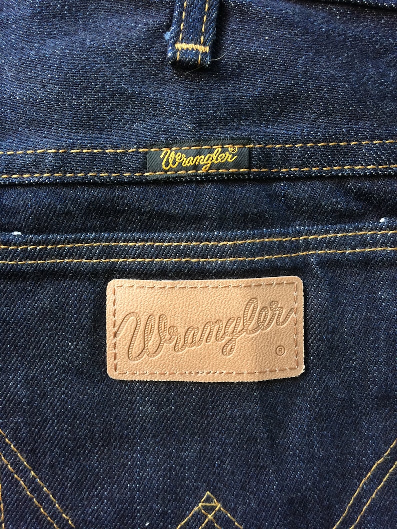 Peut inclure: Gros plan sur une paire de jeans en denim bleu fonc&eacute; avec un patch en cuir brun sur lequel est inscrit "Wrangler" en &eacute;criture cursive. Le jean a des d&eacute;tails de couture jaunes.