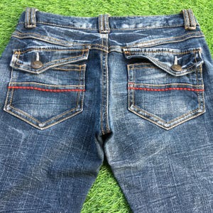 May include: Close-up of the back of blue denim jeans. The jeans have two back pockets with button closures and red stitching. The jeans have a faded, worn look.