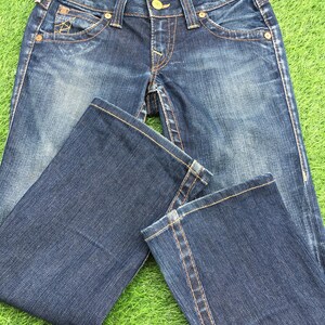 May include: Dark wash denim jeans with a classic five-pocket design. The jeans feature a button and zipper closure, with decorative stitching in a contrasting gold color. The jeans are laid out on a green surface.