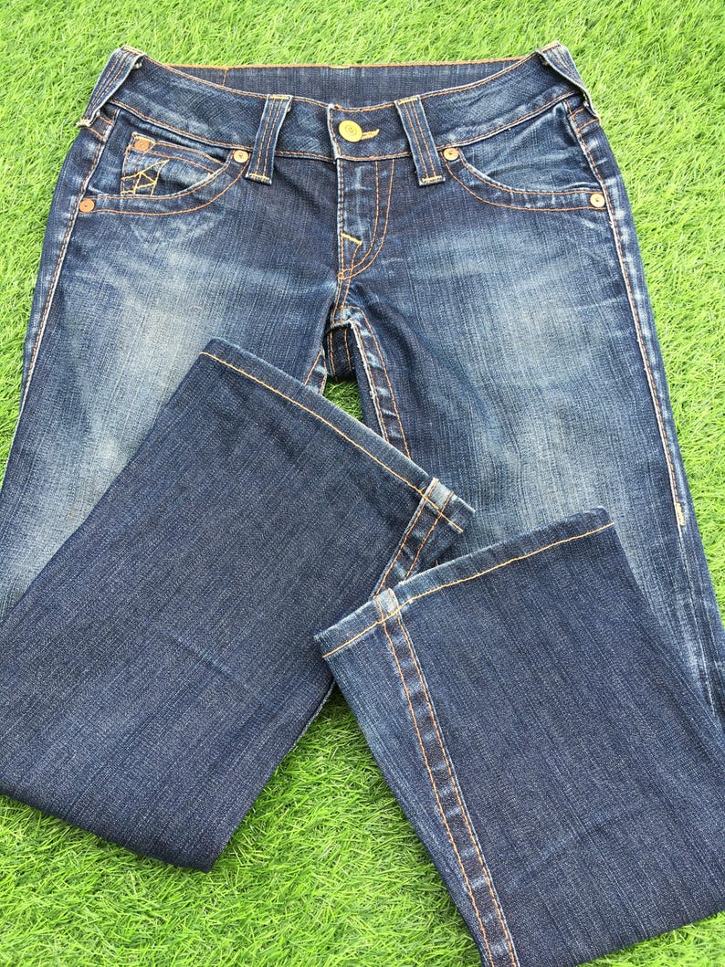 May include: Dark wash denim jeans with a classic five-pocket design. The jeans feature a button and zipper closure, with decorative stitching in a contrasting gold color. The jeans are laid out on a green surface.