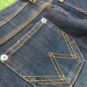 May include: Close-up of dark blue denim jeans with yellow stitching. The jeans feature a unique pocket design with a decorative stitch pattern. The back pockets have metal rivets and a leather patch.
