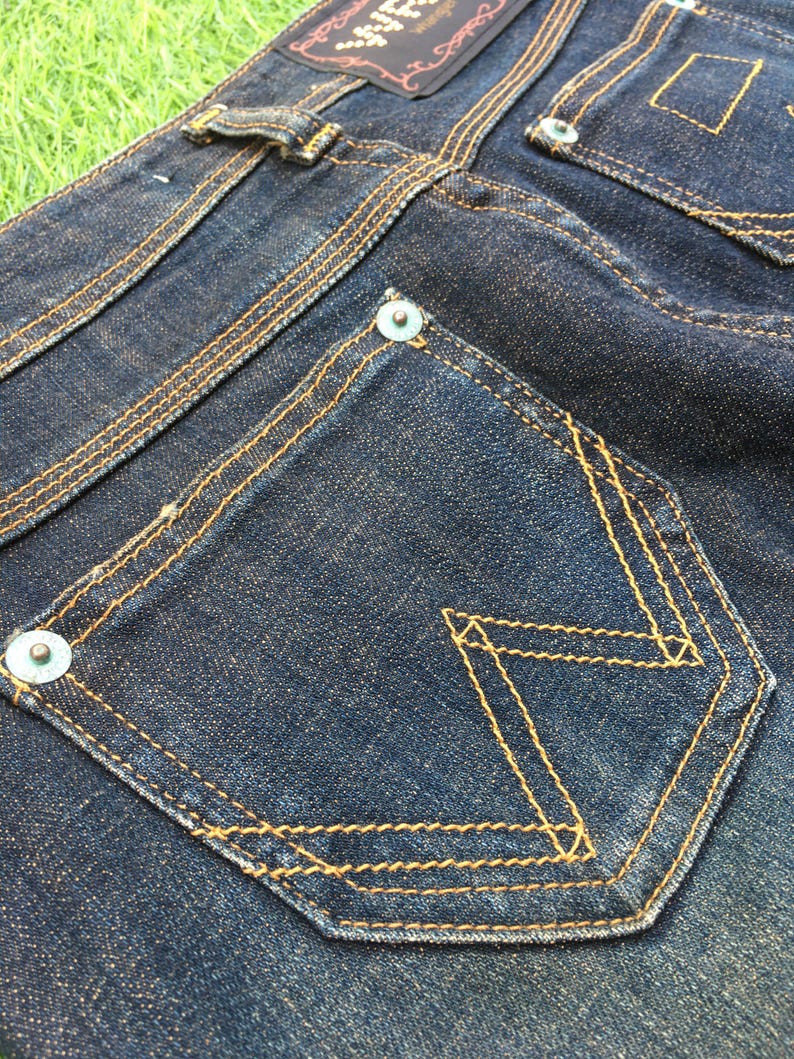 May include: Close-up of dark blue denim jeans with yellow stitching. The jeans feature a unique pocket design with a decorative stitch pattern. The back pockets have metal rivets and a leather patch.