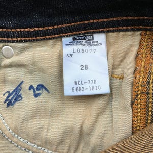 May include: Close-up of the inside of dark blue denim jeans. A white tag reads "Wrangler" and includes the size 28. The jeans have a light brown interior and a silver button.