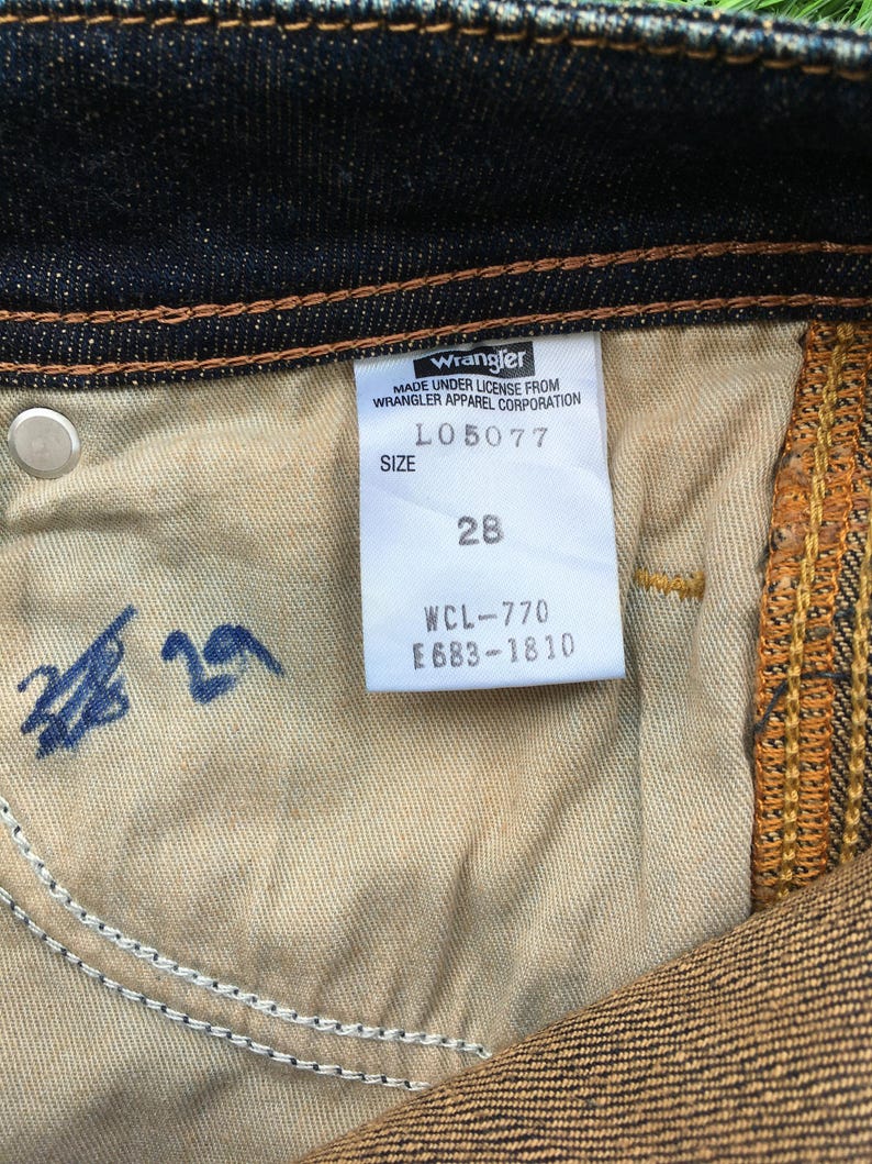 May include: Close-up of the inside of dark blue denim jeans. A white tag reads "Wrangler" and includes the size 28. The jeans have a light brown interior and a silver button.