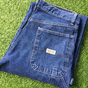 May include: A pair of dark blue denim carpenter jeans with a large pocket on the right leg. The pocket has a brown label with the text "Wrangler Jeans Co."