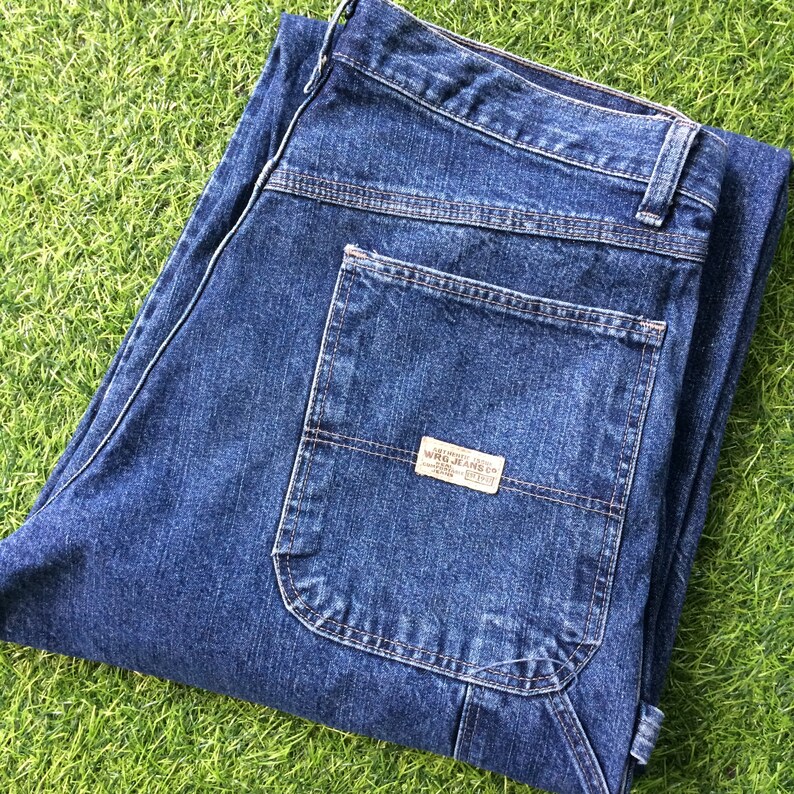 May include: A pair of dark blue denim carpenter jeans with a large pocket on the right leg. The pocket has a brown label with the text "Wrangler Jeans Co."
