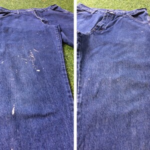 May include: A pair of blue denim carpenter jeans with white paint splatters on the front. The jeans are laid flat on a green surface.