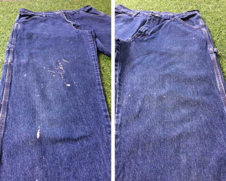 May include: A pair of blue denim carpenter jeans with white paint splatters on the front. The jeans are laid flat on a green surface.