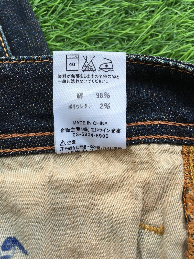 May include: Close-up of a denim jeans label with Japanese text and care symbols. The label indicates the fabric composition: 98% cotton and 2% polyurethane. The label also includes the text "MADE IN CHINA".