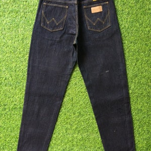Peut inclure: Un jean en denim bleu fonc&eacute; avec un &eacute;cusson en cuir marron sur la poche arri&egrave;re qui dit "Wrangler".