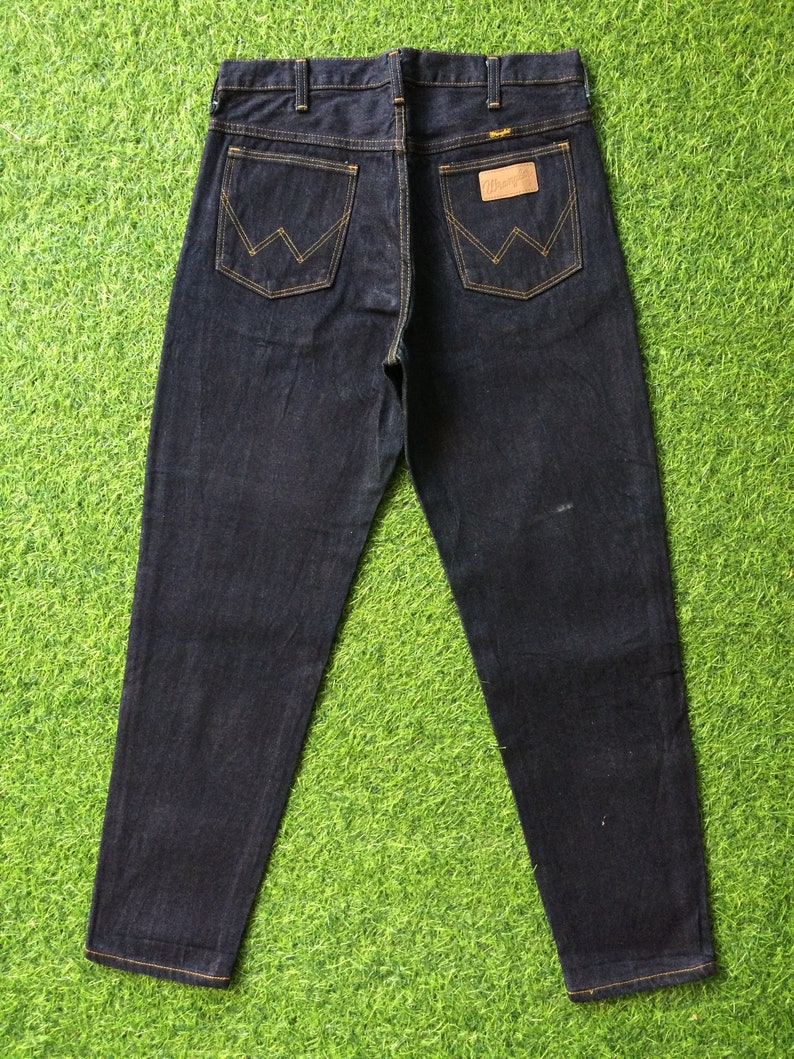 Peut inclure: Un jean en denim bleu fonc&eacute; avec un &eacute;cusson en cuir marron sur la poche arri&egrave;re qui dit "Wrangler".