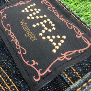 May include: Close-up of a dark denim jeans label with the brand name "Wrangler" in gold lettering. The label is black with gold-colored studs forming the word "Wrangler". A decorative pink border surrounds the label. The jeans have gold stitching.