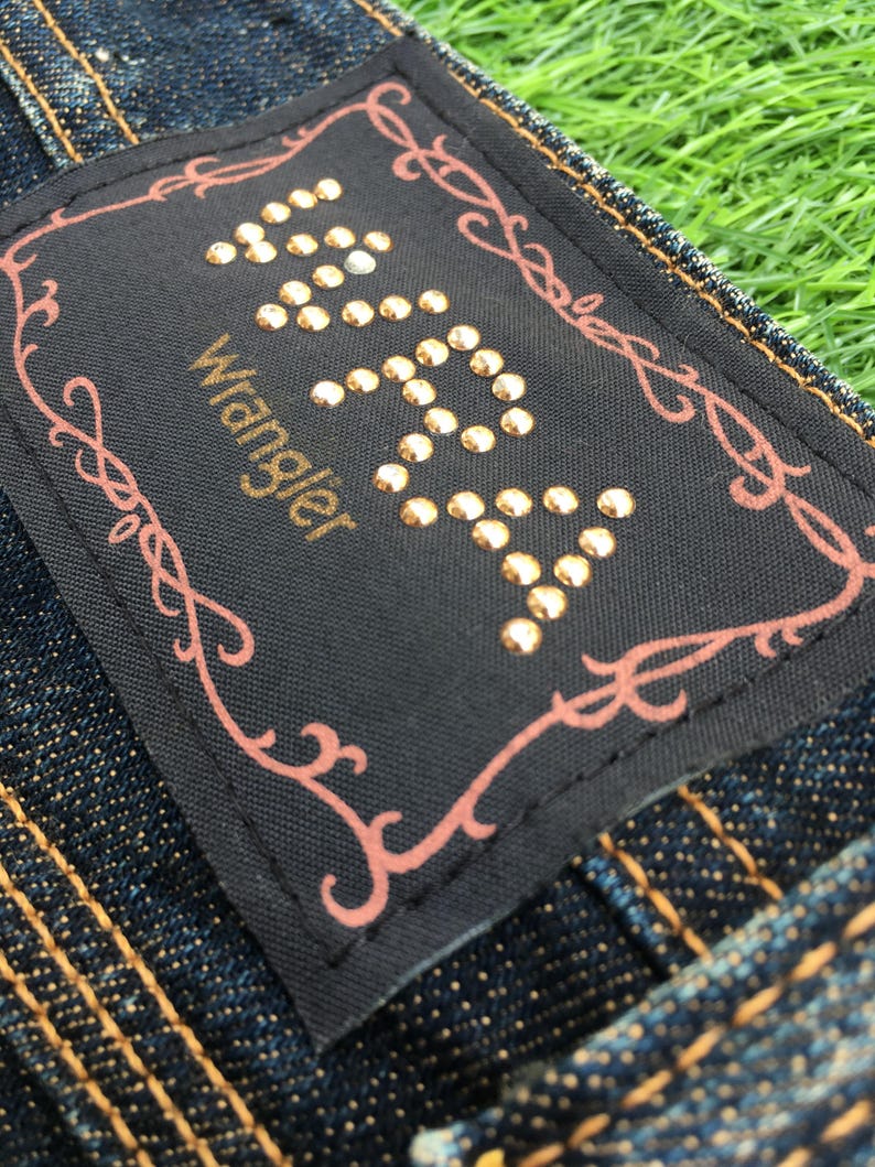 May include: Close-up of a dark denim jeans label with the brand name "Wrangler" in gold lettering. The label is black with gold-colored studs forming the word "Wrangler". A decorative pink border surrounds the label. The jeans have gold stitching.