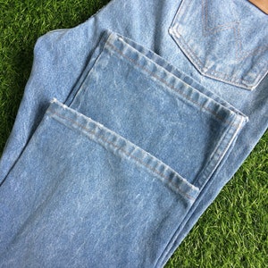 May include: A pair of light blue denim jeans with a brown leather Wrangler label. The jeans are folded in half and have a distressed look.