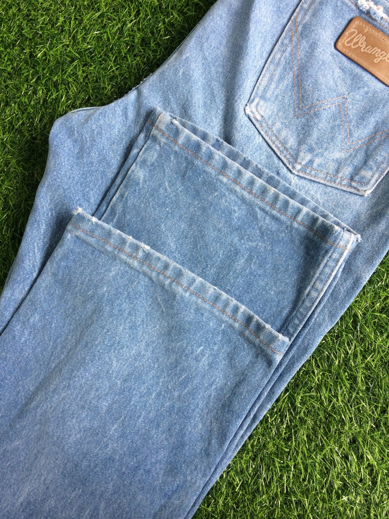 May include: A pair of light blue denim jeans with a brown leather Wrangler label. The jeans are folded in half and have a distressed look.