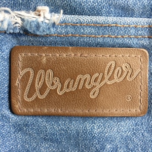 May include: A close-up of a brown leather patch with the word "Wrangler" stitched in white thread. The patch is sewn onto a blue denim fabric.
