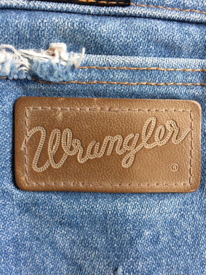 May include: A close-up of a brown leather patch with the word "Wrangler" stitched in white thread. The patch is sewn onto a blue denim fabric.