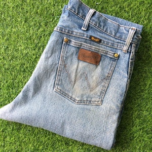 May include: A pair of light blue denim jeans with a brown leather Wrangler patch on the back pocket. The jeans have a faded look and are made of a soft, comfortable material.