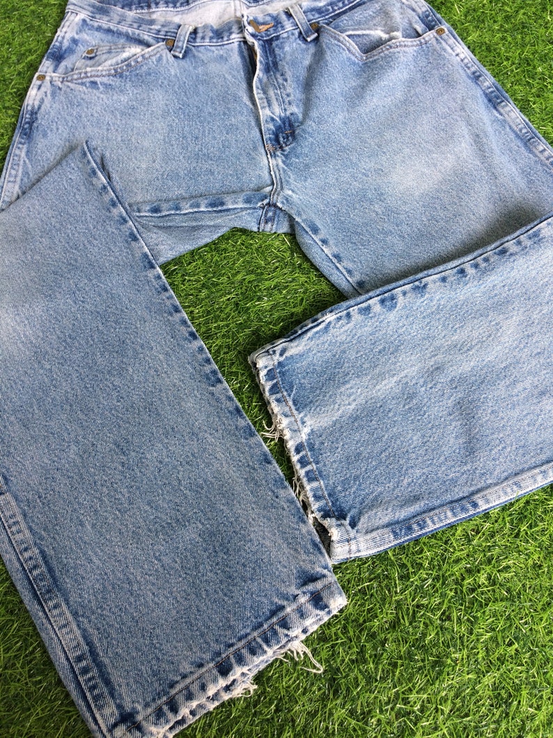 May include: A pair of light blue denim jeans with a faded wash and frayed hems. The jeans are laid out on a green grassy surface.