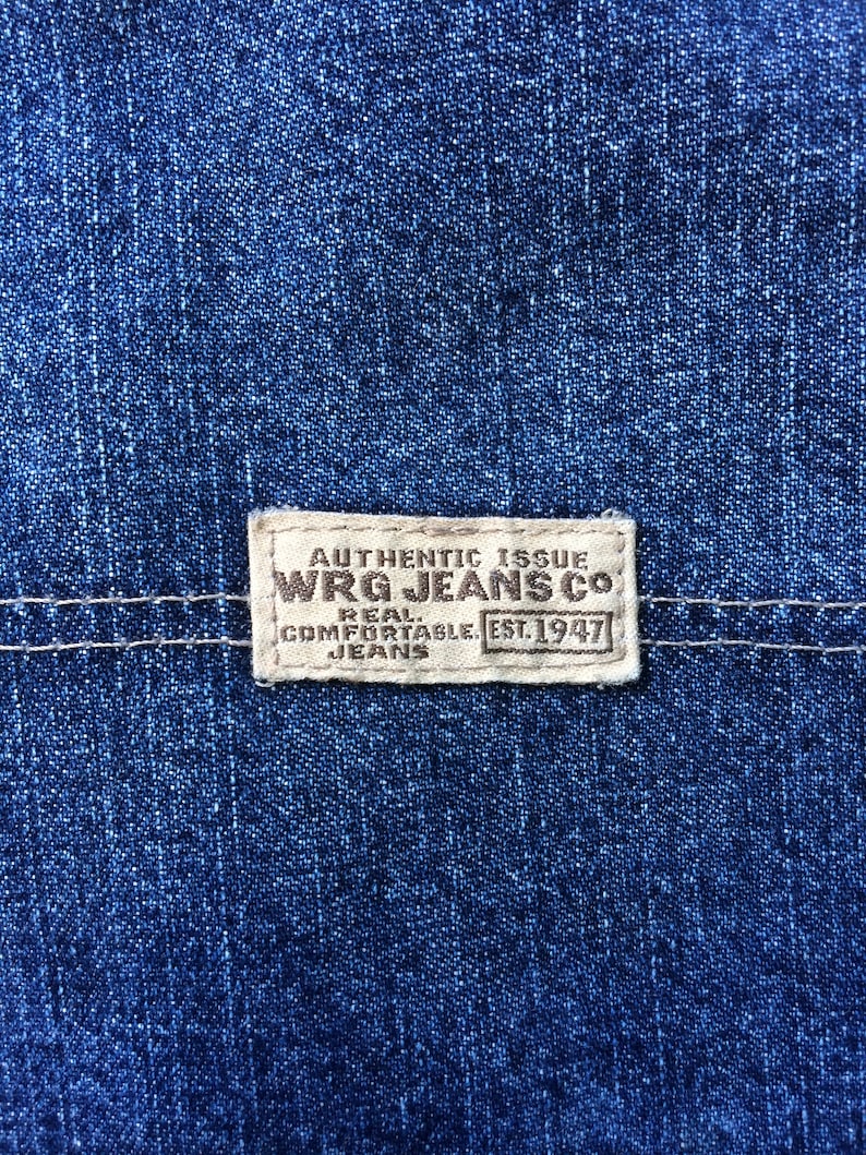 May include: A close-up of a blue denim fabric with a white label that reads "Authentic Issue WRG Jeans Co. Real Comfortable Jeans Est. 1947".