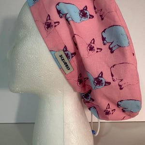 May include: A pink scrub cap with a repeating pattern of light blue and dark purple cat illustrations. The cap has a white drawstring and a small tag that reads "SCRUBBED".