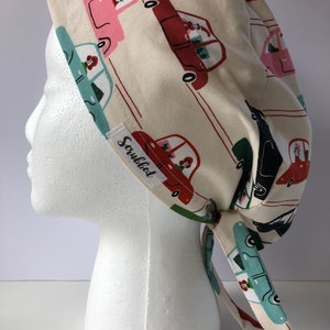 Puede incluir: Un gorro de quirófano color crema con un estampado repetido de coches antiguos en rojo, rosa, turquesa y negro. Los coches tienen detalles florales. El gorro tiene lazos y una pequeña etiqueta que dice "Scrubbed".